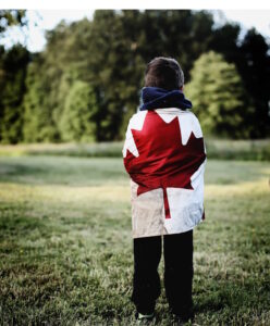 Child with his back to us, standing in a park with the Canadian flag draped over his shoulders