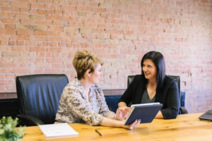 Two women talking while sitting at office table in front of brick wall