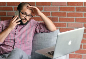 Man staring at computer screen while holding his phone. Other hand is on his forehead; he looks tired, possibly overwhelmed.